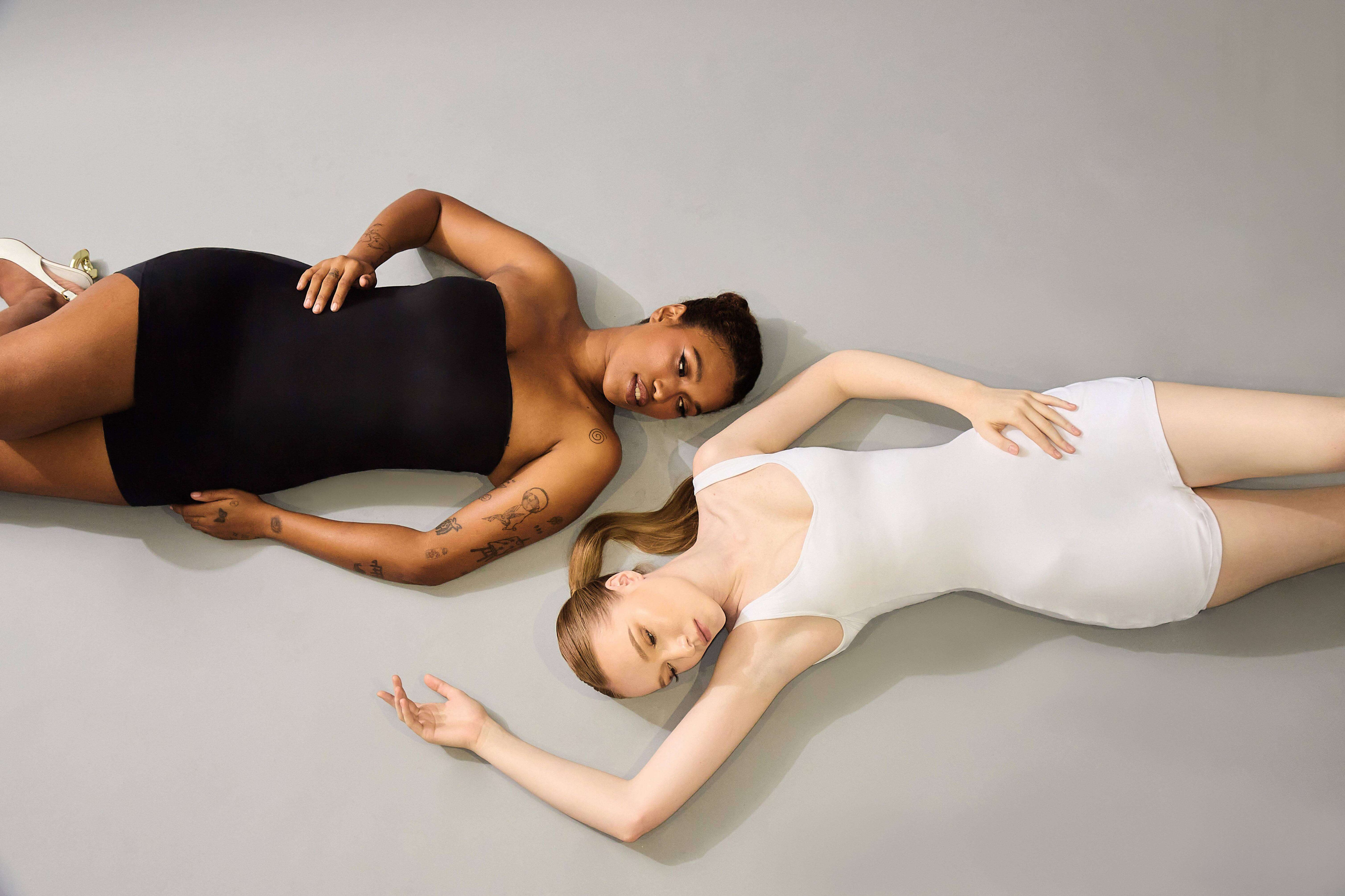 Two women lying on a gray surface, one in a black bodysuit and the other in a white bodysuit.